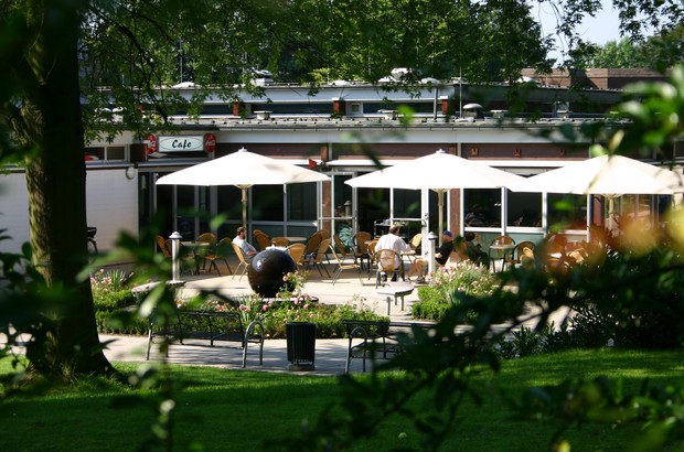 Auf einer Terrasse mit drei großen Sonnenschirmen sitzen Eineige Personen an Tische. Im Vordergrund grüner Bewuchs.